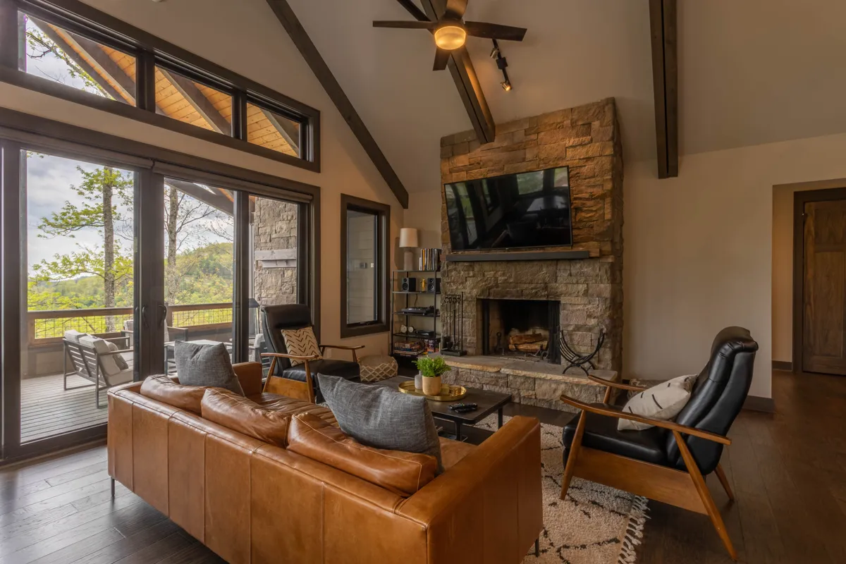 A warm, inviting living area is shown with a prominent stone fireplace, leather sofa, and large windows overlooking the mountains. Wooden ceiling beams add rustic charm, while the layout encourages socializing and relaxation. The space is styled for comfort and togetherness, ideal for game nights or gathering after a day outdoors.