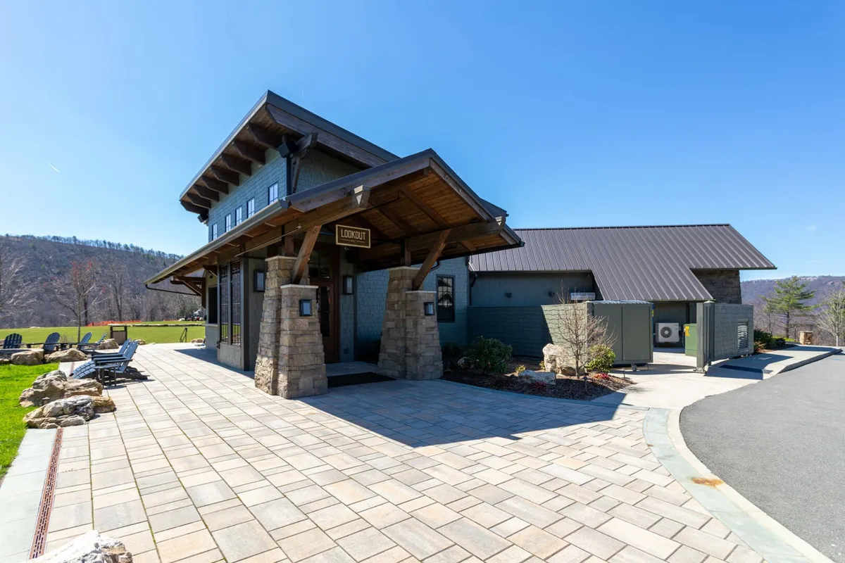 This inviting exterior shot captures the entrance of the property, featuring an awning supported by sturdy stone pillars and a paved walkway leading up to it. The scene is set against a serene mountain backdrop, with the entrance serving as the welcoming focal point.