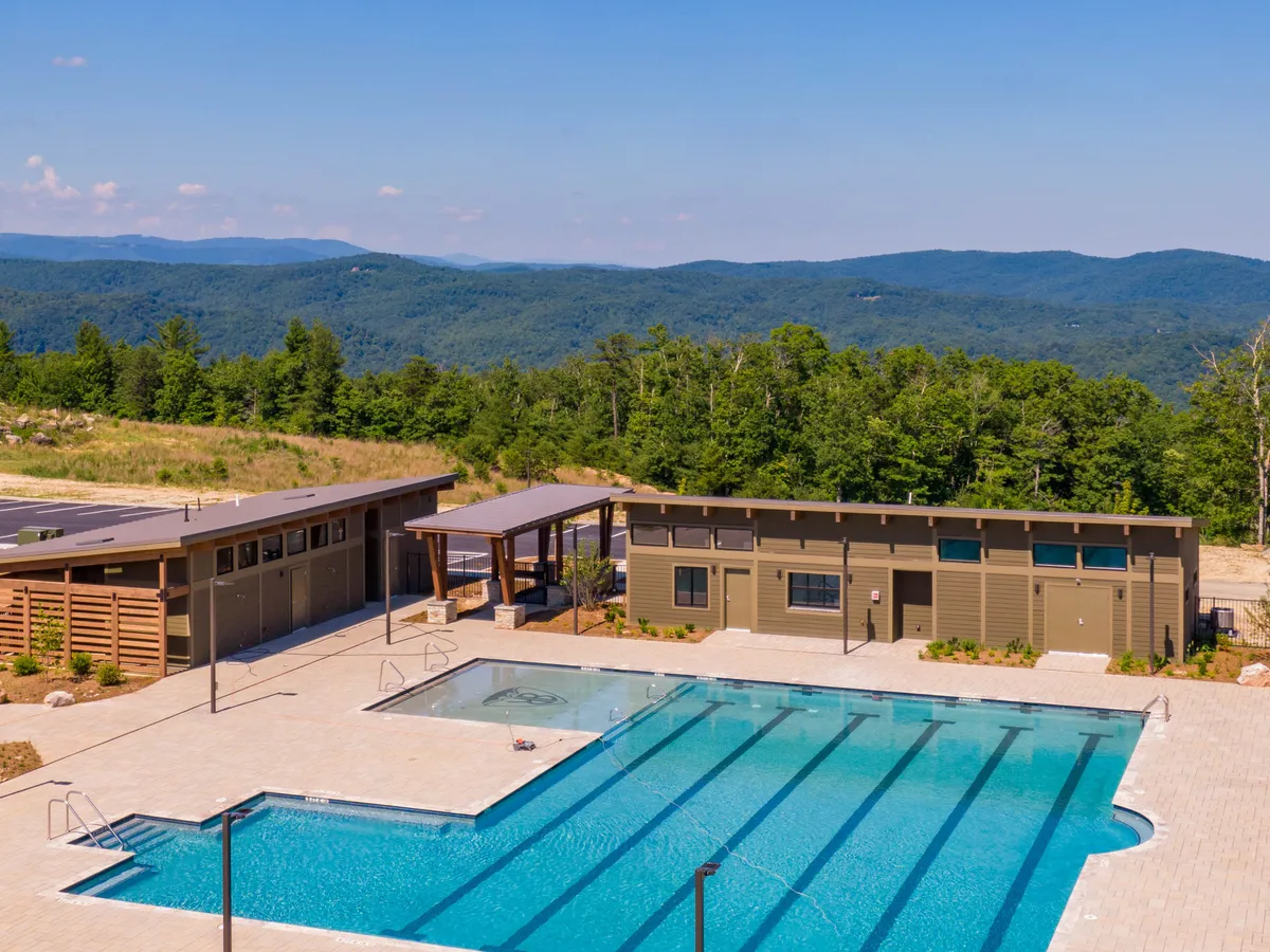 An inviting outdoor pool area with poolside cabanas, lush landscaping, and distant mountain hills. The scene includes people swimming, sunbathing, and socializing, suggesting relaxation and seasonal enjoyment. The relaxed mood and sunset light add to the feeling of a summer retreat.