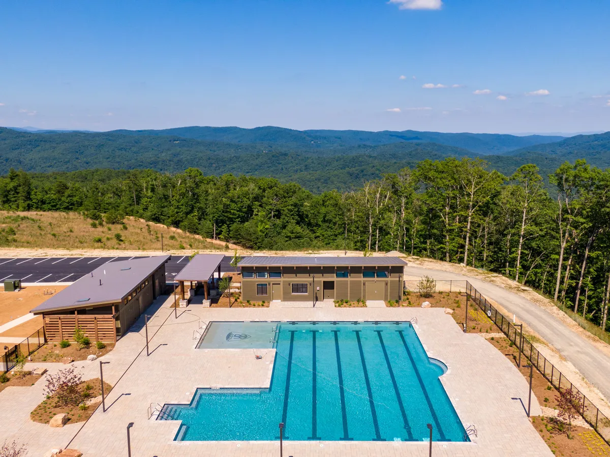 A vibrant, sunlit swimming pool is surrounded by mountains and includes a recreational area with a poolside lounge and a community facility building in the background. The area is lively, designed for social gatherings and relaxation, with children and families enjoying the space.
