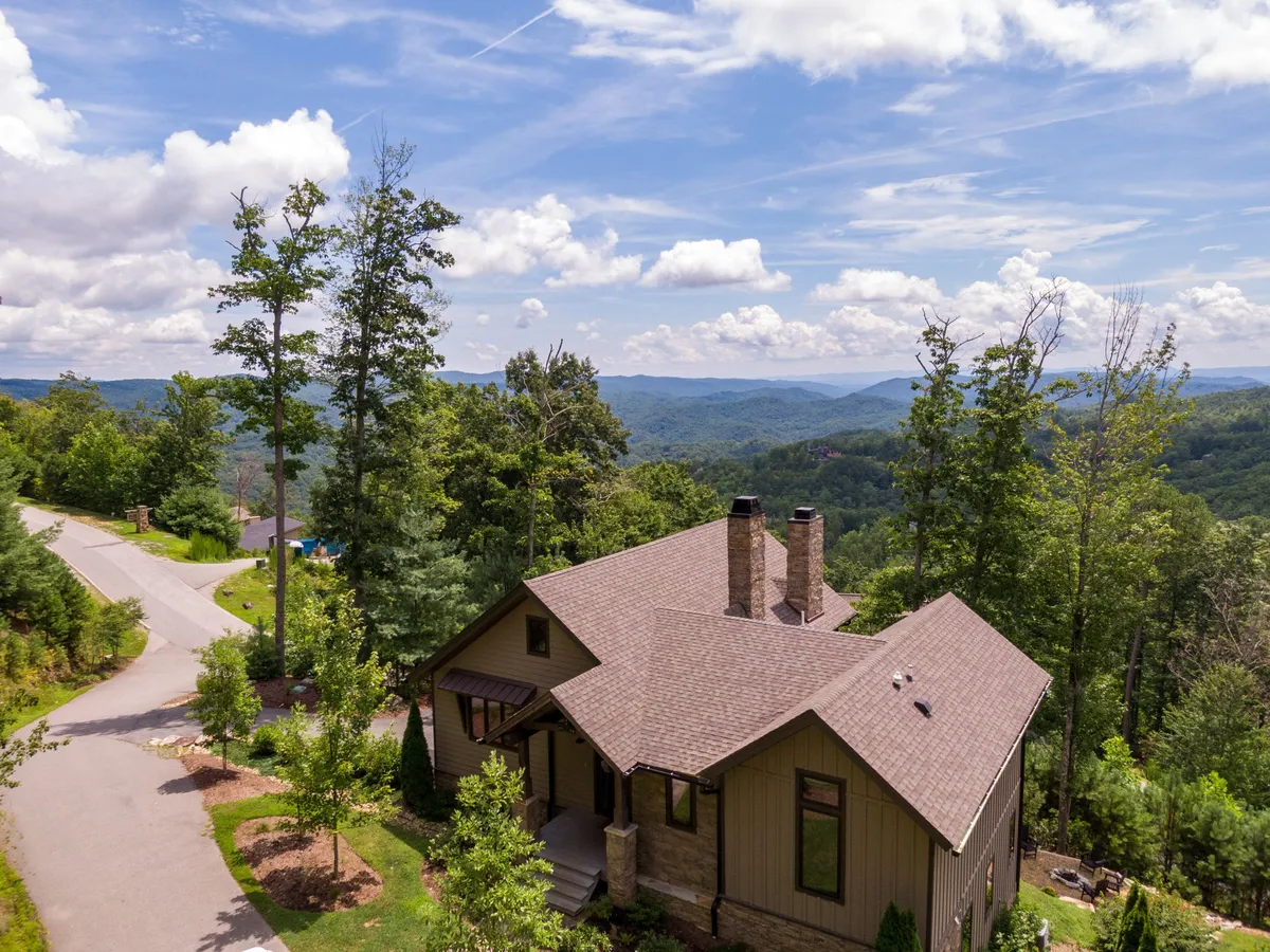 The image features the striking exterior of a contemporary mountain home nestled among lush trees, with panoramic views stretching across the Blue Ridge Mountains. The tranquil setting bathes in the warm glow of sunset, inviting guests to relax and soak in nature’s beauty. The house appears welcoming and private, promising an immersive mountain escape.