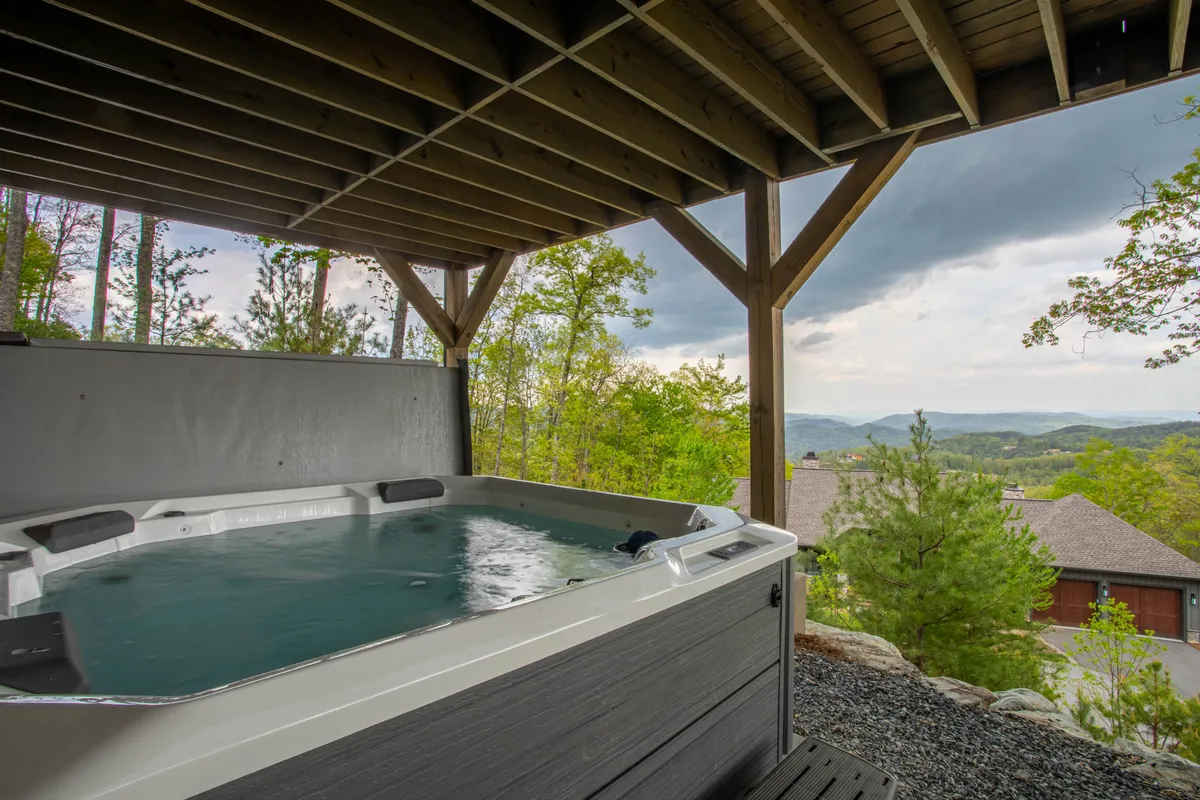 An inviting outdoor hot tub sits atop a wooden deck, overlooking a sweeping mountain vista. The setting is surrounded by natural greenery and features a sturdy wooden structure framing the space. The hot tub is positioned to maximize the panoramic view, perfect for relaxing after a day of adventure.