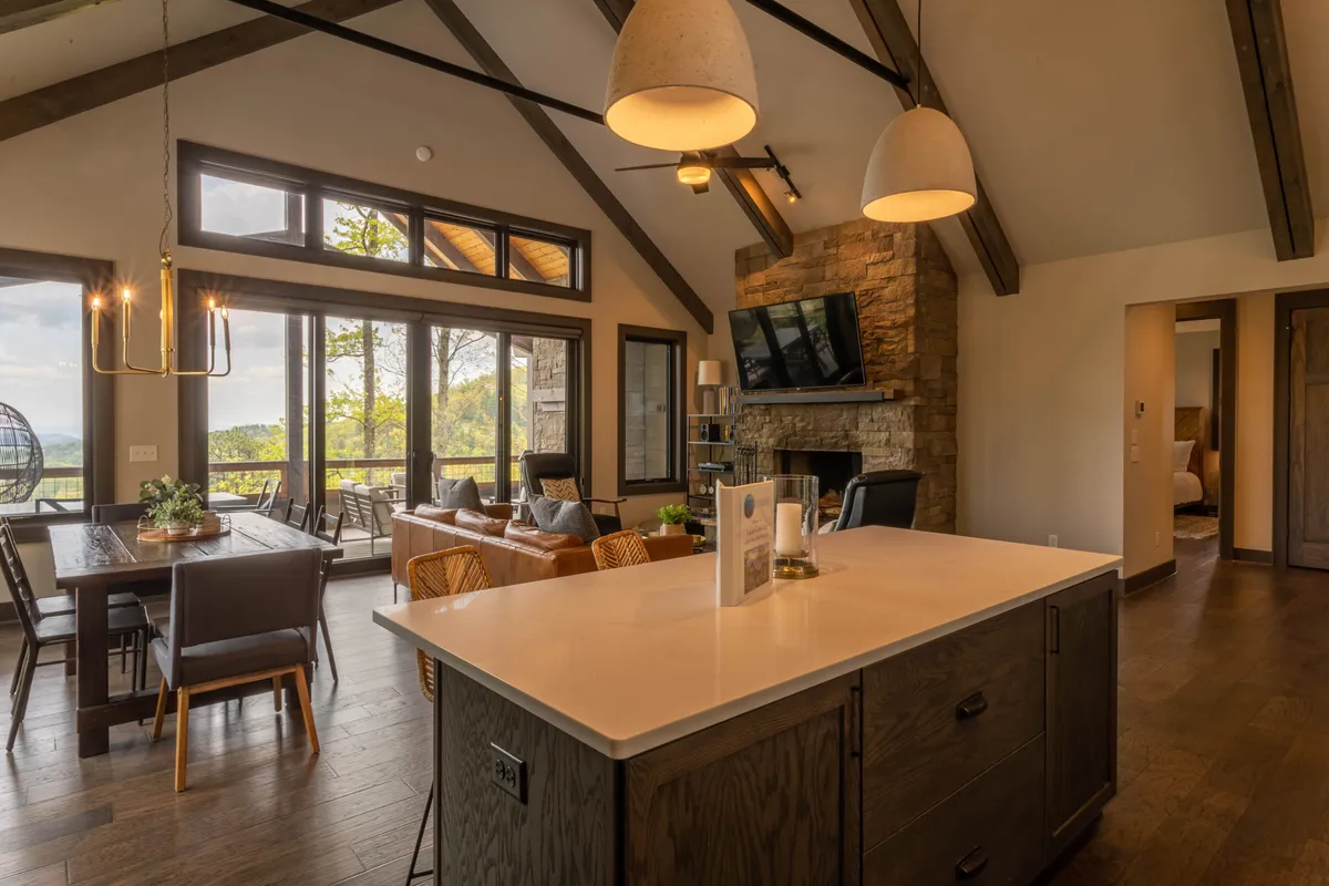 This image captures an open-concept great room that flows seamlessly into a modern kitchen and dining area. Expansive windows offer a panoramic view of the Blue Ridge mountains, allowing ample natural light to fill the space. The inviting stone fireplace anchors the living area, complemented by plush leather seating and a quartz-topped island at the center. The dining table is positioned to maximize the view, with seating for six and additional bar stools at the island.
