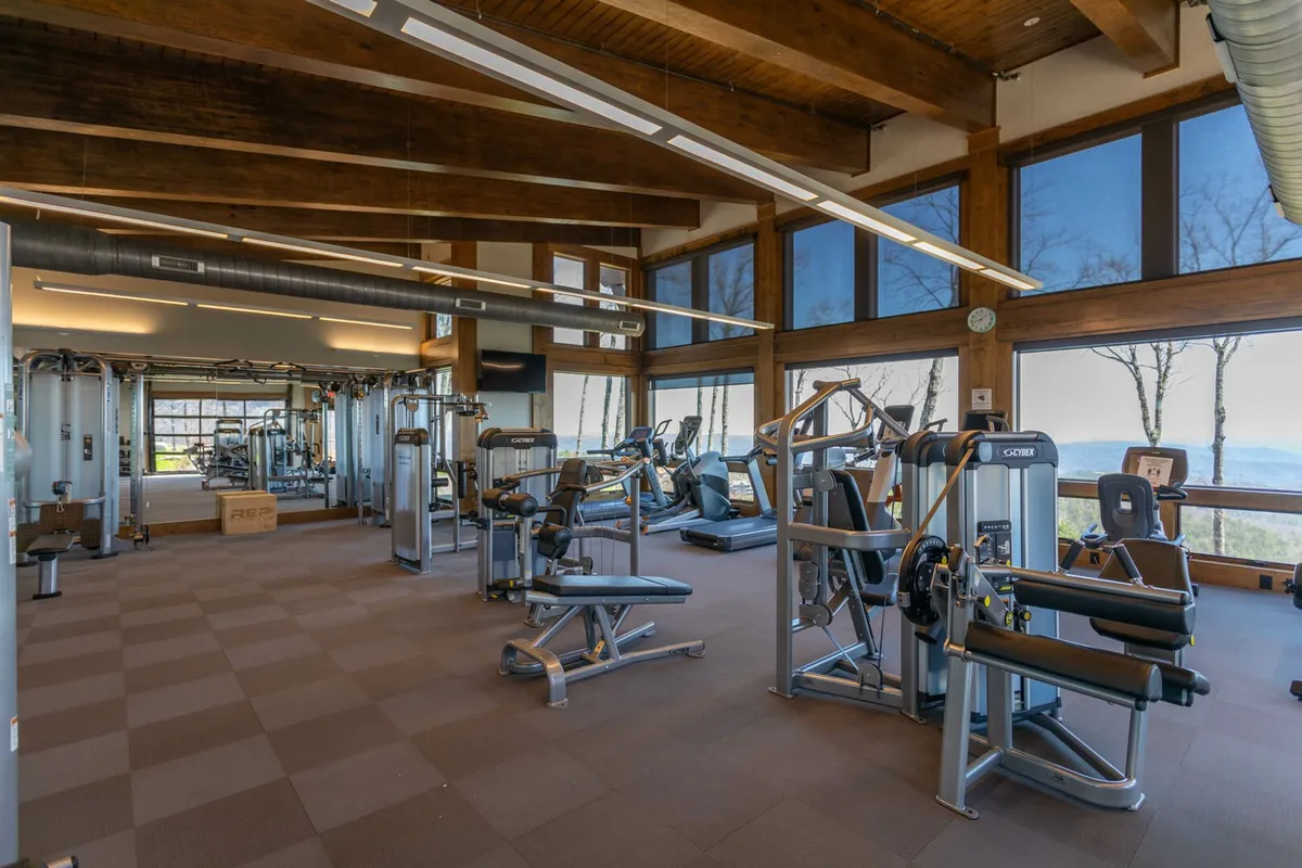 A modern interior fitness center with exercise machines, large windows revealing mountain views, and exposed wooden beams. The space is filled with natural light, creating an inviting atmosphere for both individual workouts and group fitness activities.