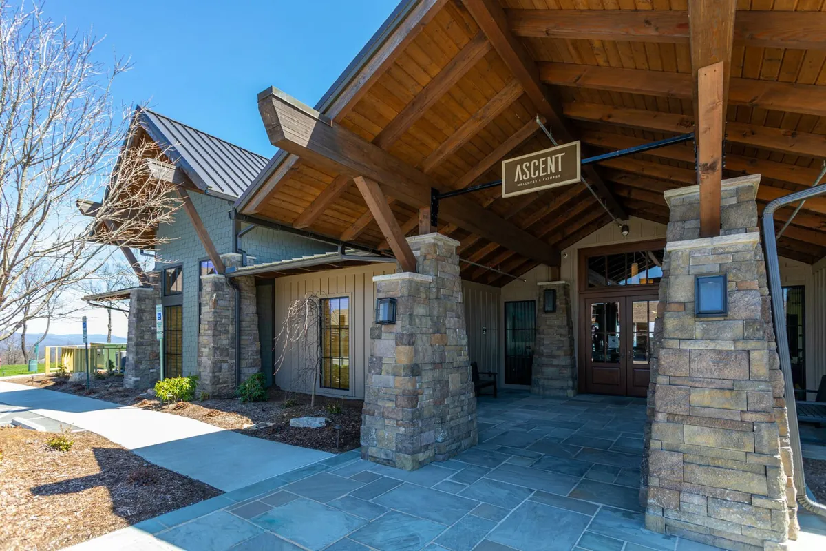 This inviting exterior image focuses on the Ascent Wellness & Fitness Center's entrance, showcasing wooden beams, stone pillars, and an 'ASCENT' sign. The surrounding landscaped area hints at the broader Blue Ridge Mountain Club amenities and the vibrant community life available to guests.