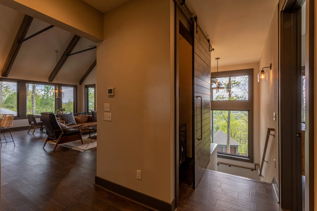A welcoming seating area sits beside large windows that reveal lush greenery outside. Modern, comfortable furniture and a sliding barn door give the space a contemporary mountain feel. The view and layout suggest a perfect environment for relaxed mornings, evenings by the fire, or game nights with friends.