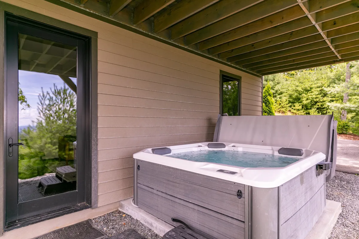 This image features a private hot tub nestled on an outdoor patio, surrounded by lush greenery. The sliding glass door hints at a seamless transition between indoor comfort and the natural world outside. The setup invites relaxation and soaking in the tranquil environment, with views of the landscape offering a sense of peace and privacy.