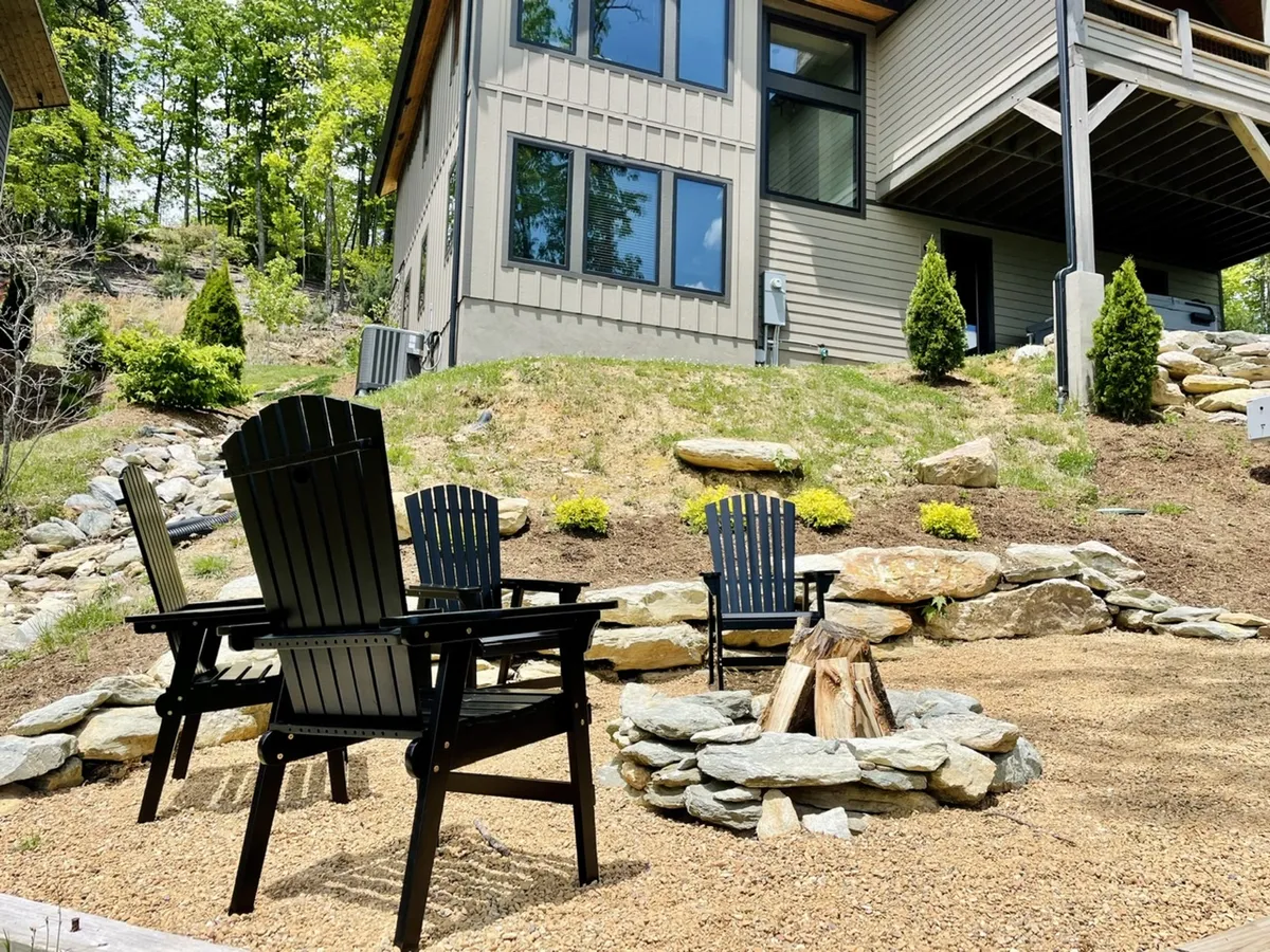 A landscaped fire pit area with black Adirondack chairs arranged in a circle, facing the house and surrounded by greenery. The mood is relaxed, inviting guests to gather around the fire for stories and s'mores.